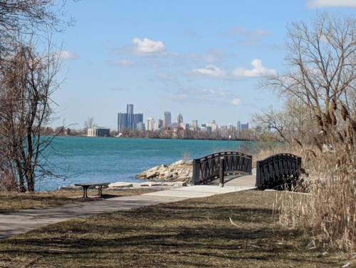 A scenic view of a city skyline across a blue lake, with a bridge and pathway in the foreground.