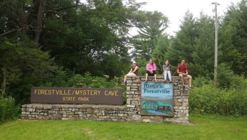 Four people sit on a stone sign for Forestville/Mystery Cave State Park, surrounded by greenery.