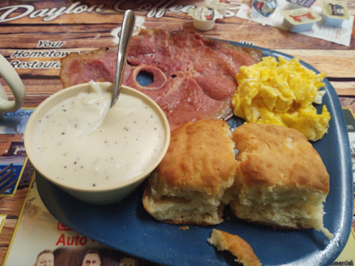 A plate of breakfast featuring ham, scrambled eggs, biscuits, and a bowl of creamy gravy.