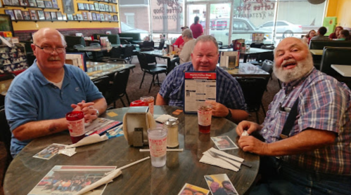 Three men sit at a restaurant table, smiling and holding a menu, with drinks and photos scattered around.