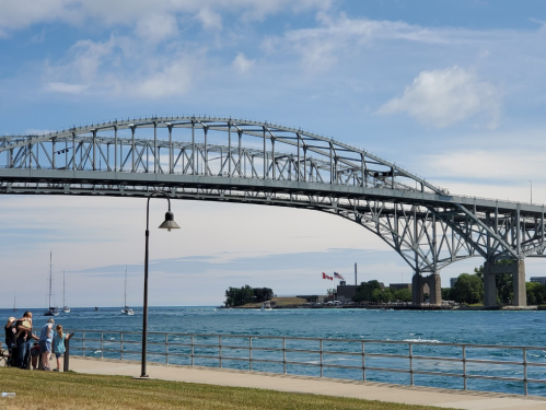 A large bridge spans over a blue waterway, with people gathered on the shore and boats in the distance under a clear sky.