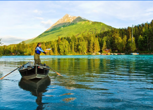 A person fishing from a boat on a calm lake, surrounded by lush trees and a mountain in the background.