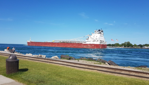 A large cargo ship sails through a blue waterway under a clear sky, with people sitting nearby on the shore.