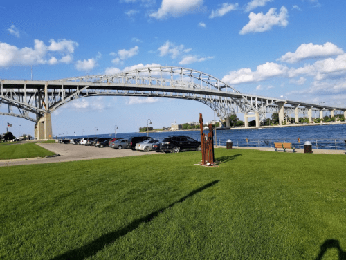 A bridge spans over a river, with green grass, parked cars, and a blue sky dotted with clouds in the background.