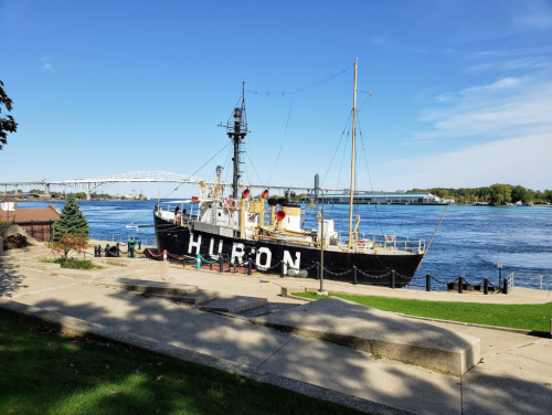 A black ship named "HURON" docked by a river, with a bridge in the background and clear blue skies above.