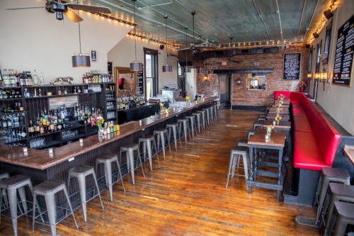 A spacious bar interior with wooden floors, a long counter, red seating, and shelves of drinks against a brick wall.