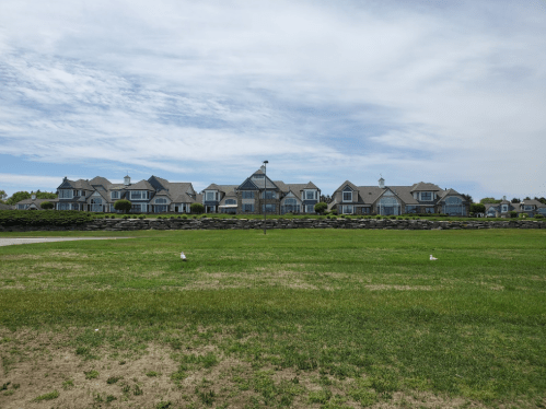 A row of large houses with gray roofs along a grassy area under a partly cloudy sky.