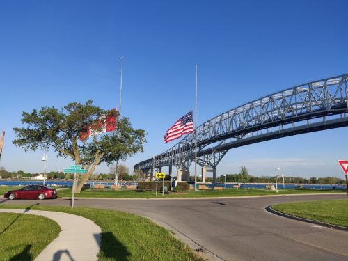 A blue bridge spans a river, with flags at half-mast and a red car parked nearby. Clear blue sky above.