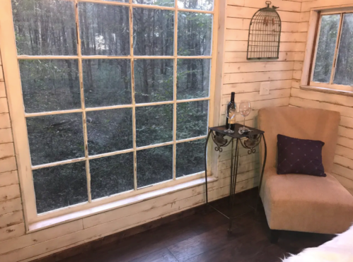 Cozy interior with a large window overlooking a forest, featuring a chair, a small table, and a bottle of wine.