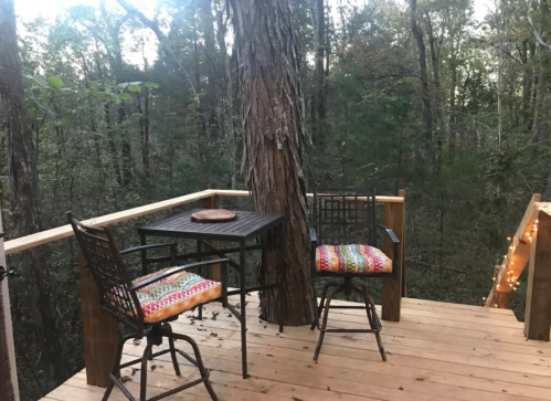 A cozy wooden deck in the woods with two chairs and a small table, surrounded by trees and soft lighting.