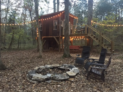A cozy treehouse with string lights, surrounded by trees and a fire pit, with two chairs nearby on a leaf-covered ground.