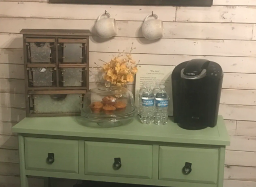 A green table with a wooden drawer unit, water bottles, a coffee maker, and a cake stand with muffins, set against a wooden wall.