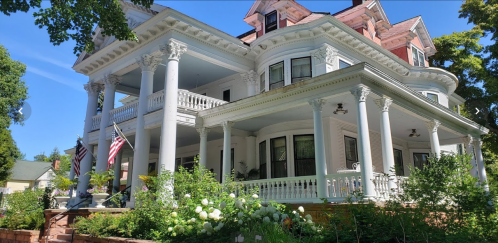 A large, elegant Victorian-style house with white columns, surrounded by greenery and American flags.