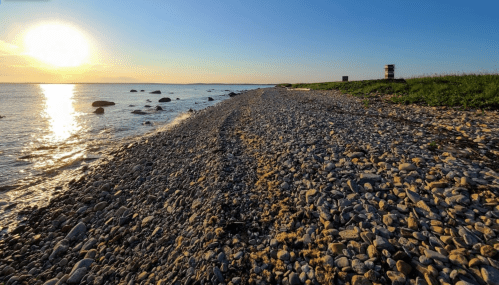 A rocky shoreline at sunset, with the sun reflecting on the water and a distant lookout tower.
