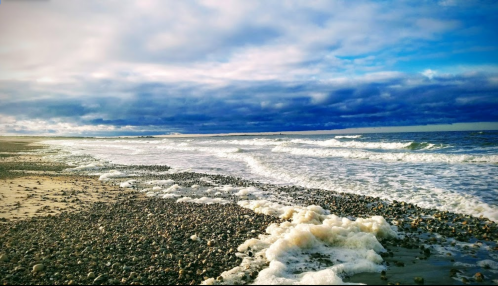 A serene beach scene with gentle waves, pebbles on the shore, and a cloudy blue sky above.
