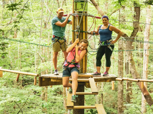 Three people in harnesses on a treetop platform, surrounded by trees, preparing for a ropes course adventure.