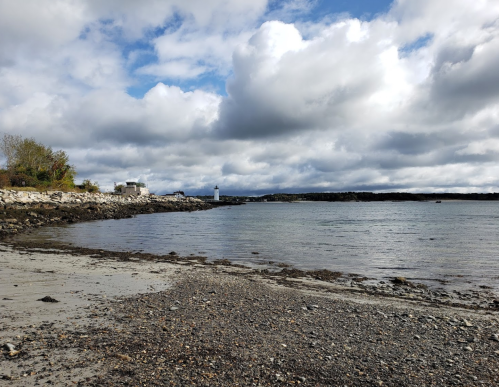 A rocky beach with a lighthouse in the distance under a cloudy sky. Calm water reflects the clouds.