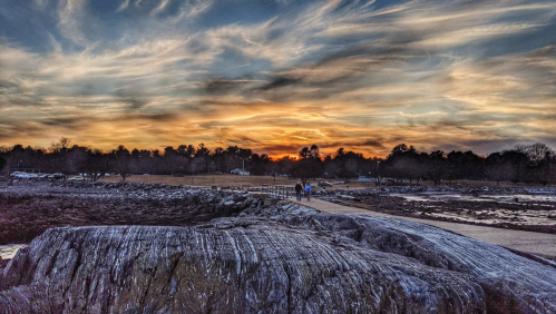 A scenic view of a rocky shoreline at sunset, with people walking along a path and dramatic clouds in the sky.