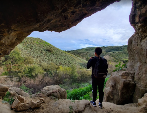 A person stands in a cave opening, gazing at a lush green landscape under a cloudy sky.
