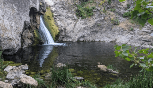 A serene waterfall cascades into a calm pool surrounded by rocky cliffs and lush greenery.