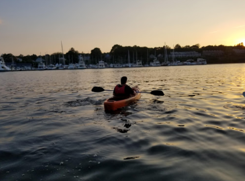 A person kayaking on calm water during sunset, with boats docked in the background.