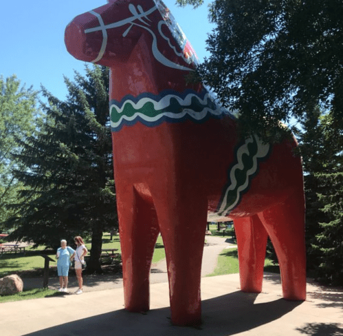 A large red Dala horse statue stands in a park, surrounded by trees, with two people walking nearby.