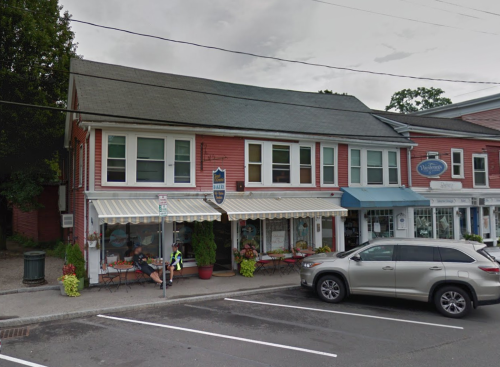 A pink building with white awnings, featuring a café and shops, parked cars in front, and a person sitting outside.