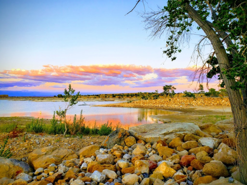 A serene lakeside scene at sunset, featuring colorful clouds, rocky shorelines, and lush greenery.