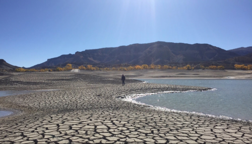 A person walks on cracked earth near a small body of water, with mountains and autumn trees in the background under a clear sky.