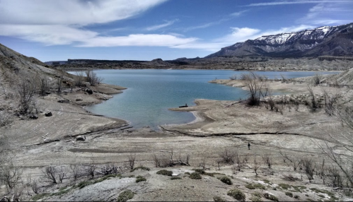 A serene lake surrounded by dry land and mountains under a clear blue sky, with sparse vegetation along the shore.