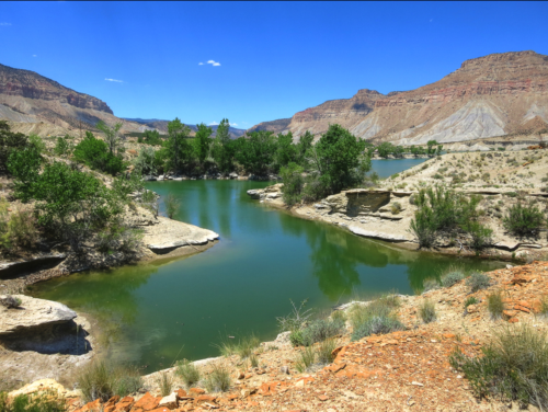 A serene river flows through a rocky landscape, surrounded by green trees and mountains under a clear blue sky.
