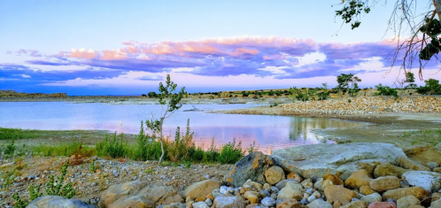 A serene lakeside view at sunset, with colorful clouds reflecting on the water and rocky shores lined with greenery.