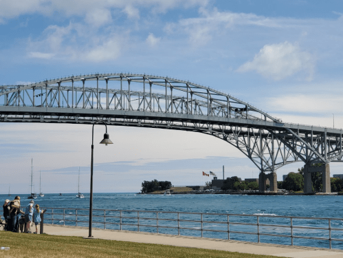 A large steel bridge spans over a blue waterway, with people gathered on the shore and sailboats in the distance.