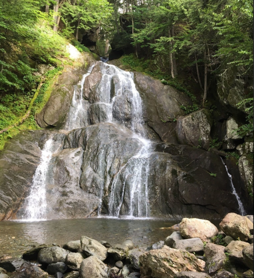 A serene waterfall cascading down rocky terrain, surrounded by lush green trees and a calm pool at the base.