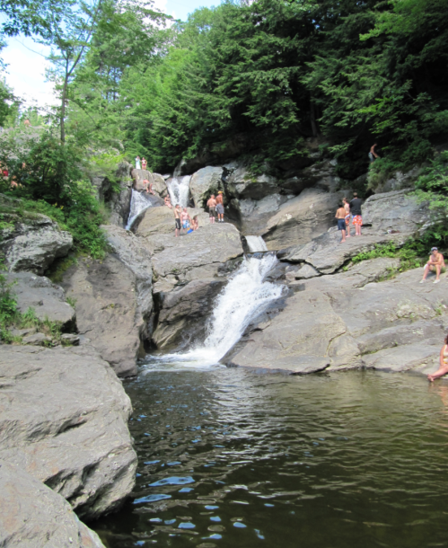 A scenic waterfall surrounded by rocks and trees, with people swimming and relaxing by the water.