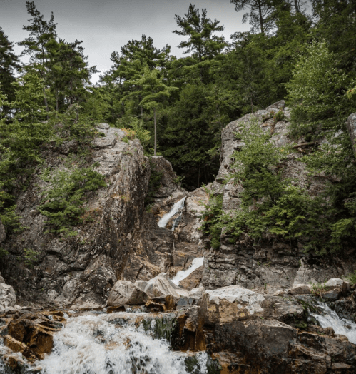 A rocky landscape with a waterfall cascading down, surrounded by lush green trees and rugged cliffs.