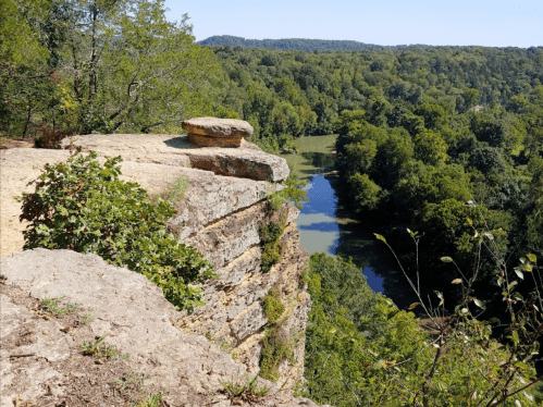A rocky cliff overlooks a lush green valley and a winding river under a clear blue sky.