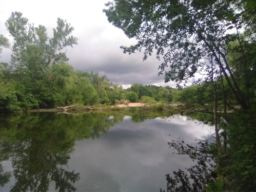 A serene river scene surrounded by lush greenery and cloudy skies, reflecting the landscape in calm waters.