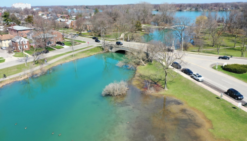 Aerial view of a vibrant blue pond with a bridge, surrounded by trees and houses, on a sunny day.