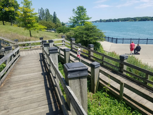 A wooden walkway leads to a waterfront, with a child walking and two people in the distance near the water.