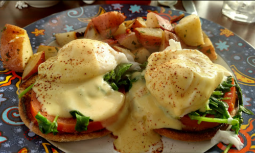 A colorful plate with two eggs Benedict on toasted English muffins, topped with hollandaise sauce and served with seasoned potatoes.
