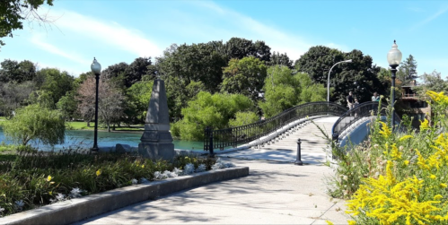 A scenic park view featuring a bridge over a pond, surrounded by greenery and flowers under a clear blue sky.