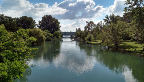 A serene river scene with lush greenery, a bridge in the distance, and fluffy clouds reflecting in the water.