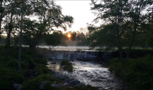 A serene river flows over a small waterfall, framed by trees and a rising sun in the background.