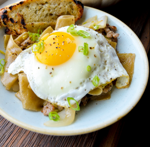 A bowl of noodles topped with a fried egg, green onions, and a slice of toasted bread.