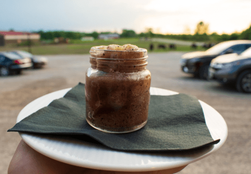 A jar of dessert on a plate, held outdoors with a scenic background of cars and a field.
