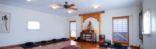 A serene meditation room with cushions, a Buddha statue, and wooden accents, featuring natural light from windows.