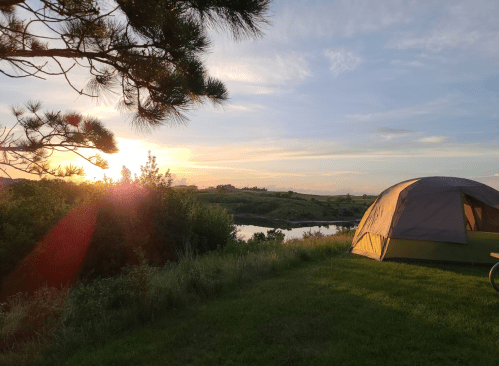 A tent by a river at sunset, surrounded by greenery and trees, with a colorful sky in the background.