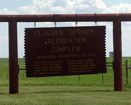 Sign for Clausen Springs Recreation Complex, listing various organizations involved in its development. Green fields in background.