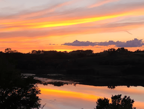 A vibrant sunset with orange and pink hues reflecting on a calm lake, surrounded by trees and hills.